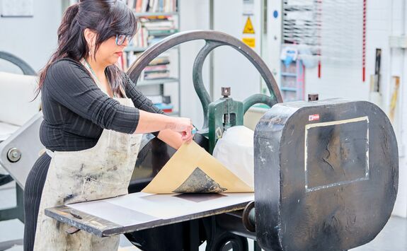 Person in an apron operating a printing press in an art studio, handling a sheet of paper. Shelves with books and tools are visible in the background.